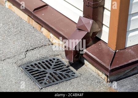 Regenwasserleitung an der Wand der Gebäudefassade mit Regenwasserableitung in das Abflusssystem mit Kanalgrill. Stockfoto