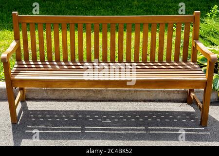 Eine hölzerne braune Bank aus Brettern steht auf einem Asphaltbelag nahe der Bordsteinkante mit einer grünen Wiese im Park. Stockfoto