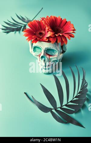 Totenkopf mit korallenfarbenen Gerbera Gänseblümchen und dunkelgrünen Palmenblättern auf kachelgrünem Hintergrund. Kreatives Konzept. Surreales Gefühl, unheimliche gruselige Atmosphäre Stockfoto