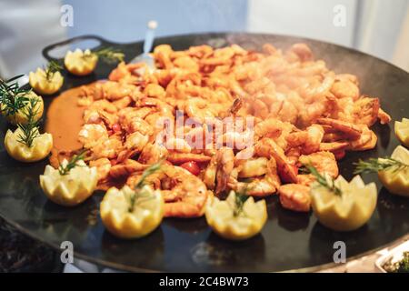 Nahaufnahme der Garnelen mit Sauce auf großer schwarzer Pfanne mit Zitronenblüten garniert verziert. Feier, Party, Geburtstag oder Hochzeit Konzept. Stockfoto