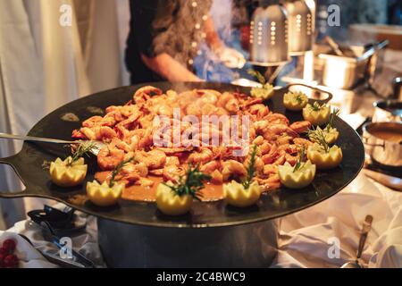 Große schwarze Pfanne mit Garnelensalat mit Sauce und mit Zitronenblüten garniert. Im Hintergrund kochen. Feier, Party, Geburtstag oder Hochzeit. Stockfoto