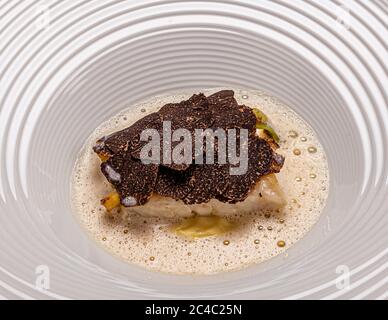 Scheiben von schwarzem Trüffel bedecken ein weißes Fischfilet in einer blassen, schaumigen Sauce, serviert auf einem tiefen weißen Teller mit gezacktem Rand. Grüne und gelbe Gemüsestücke liegen unter dem Fisch. Das Gericht gehört zum Galadinner des mit einem Michelin-Stern ausgezeichneten Kochs Klaus Erfort am 27. Februar 2017 im Hotel und Restaurant Kronenschlösschen in Hattenheim, Eltville am Rhein, Hessen, Deutschland. Das Galadinner ist Teil des 21. Rheingau Gourmet & Wein Festivals. Erfort führt das Gästehaus Klaus Erfort in Saarbrücken. Er hat seit 2008 drei Michelin-Sterne. Zu dieser Zeit bewertet ihn Gault-Millau mit 19,5 Punkten. Meister so Stockfoto