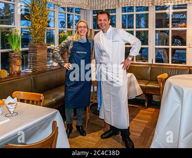 Der Michelin-Sterne-Koch Thomas Kellermann (rechts) steht mit der Journalistin Angela Berg (links) im Restaurant des Hotels und Restaurants Kronenschlösschen in Hattenheim, Eltville am Rhein, Hessen, Deutschland. Kellermann trägt eine weiße Kochjacke mit seinem aufgestickten Namen und eine weiße Schürze. Forsythienzweige und Narzissen stehen an den Gitterfenstern hinter ihnen. Thomas Kellermann ist Küchenchef des Restaurants Kastell im Schloss Burg Wernberg in der Oberpfalz, Bayern, wo er seit 2011 zwei Michelin-Sterne hält. Zuvor kochte er von 1994 bis 2000 im Tantris in München, am Stockfoto