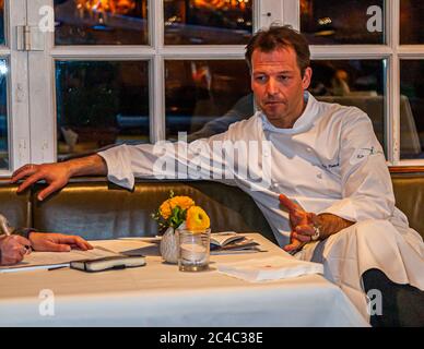 Der mit einem Michelin-Stern ausgezeichnete Koch Thomas Kellermann sitzt auf einer Lederbank im Restaurant des Hotels und Restaurants Kronenschlösschen in Hattenheim, Eltville am Rhein, Hessen, Deutschland, und gestikuliert, während er mit der Journalistin Angela Berg spricht. Er stützt seinen rechten Arm auf die Rückenlehne und hebt seine linke Hand. Angela Bergs Hände mit einem Stift, Papieren und einem Notizbuch liegen am linken Rand des Tisches. Gelbe Ranunkeln in einer Vase und ein Kerzenhalter stehen auf dem Tisch. Kellermann trägt eine weiße Kochjacke. Thomas Kellermann ist Küchenchef des Restaurants Kastell im Burg Wernberg in der Oberpfalz, Stockfoto