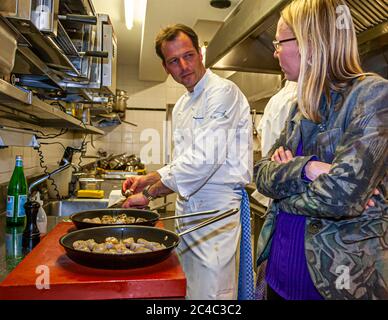 Michelin-Sterne-Koch Thomas Kellermann (links) arbeitet an zwei Pfannen mit gebratenen Jakobsmuscheln in der Küche des Hotels und Restaurants Kronenschlösschen in Hattenheim, Eltville am Rhein, Hessen, Deutschland, und schaut auf Journalistin Angela Berg (rechts). Sie steht neben ihm mit verschränkten Armen und trägt eine grau-grün gemusterte Jacke über einem lila Oberteil. Die Pfannen stehen auf einem roten Schneidebrett. Kellermann trägt eine weiße Kochjacke mit seinem Namen bestickt und ein blau-weiß kariertes Tuch auf seiner Schürze. Thomas Kellermann ist Küchenchef des Restaurants Kastell im Schloss Burg Wernberg in der Oberpfalz Stockfoto