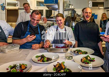 Spaß in der Küche: Köche lachen an einem Tisch mit weißer Tischdecke und angerichteten Gerichten in der Küche des Hotels und Restaurants Kronenschlösschen in Hattenheim, Eltville am Rhein, Hessen, Deutschland. Ein Koch in einer blauen Schürze hält ein Smartphone, eine junge Köchin hält ein Tablett mit roten Würfeln. Rechts lacht Gastgeberkoch Simon Stirnal in einer schwarzen Kochjacke. Dunkelrote Scheiben, orangefarbene Chips, weiße Stücke und Kräuter liegen auf den Tellern. Der mit einem Michelin-Stern ausgezeichnete Koch Simon Taxacher und sein Team haben die Gerichte angerichtet. Am Abend des 10. März 2017 kocht der mit einem Michelin-Stern ausgezeichnete Koch Simon Taxacher eine "Kulinarische Oper in fünf Gä Stockfoto