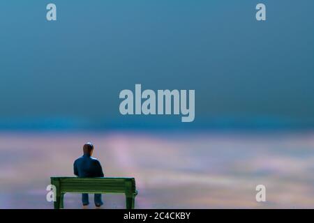 Miniatur-Figur posiert als Mann allein auf einer Bank sitzen in surrealen Landschaft, minimalistisch abstraktes Konzept Bild Stockfoto