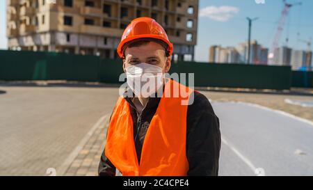 Porträt des männlichen Bauarbeiters in medizinischer Maske und Overalls auf dem Hintergrund des Hauses im Bau. Stockfoto