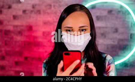 Junge schöne Frau in medizinischen Maske steht mit Handy in den Händen auf Backsteinwand mit glühenden Lampen. Porträt von charmanten weiblich Stockfoto