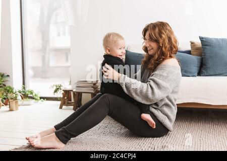 Junge lächelnde Frau, die auf dem Boden sitzt und mit ihrem kleinen, hübschen Sohn spielt. Mama mit Baby Junge freudig Zeit im Wohnzimmer zu Hause verbringen Stockfoto