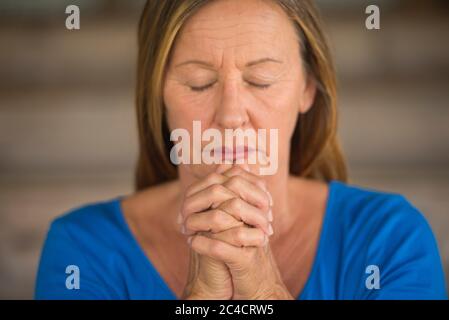 Portrtait Attraktive religiöse ältere Frau beten mit Fokus auf gefaltete Hände, nachdenklich, meditierend, geschlossene Augen, verschwommener Hintergrund. Stockfoto