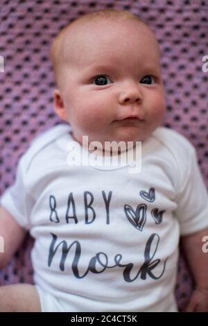 Ein neugeborener glücklicher Junge, der auf einem lila Teppich mit einer weißen Babyweste mit den Worten Baby Mork liegt. Foto von Sam Mellish Stockfoto