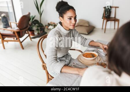 Junge schöne lächelnde afroamerikanische Frau mit dunklen lockigen Haaren sitzt am Tisch verträumt neben, während die Zeit mit Freund in Stockfoto