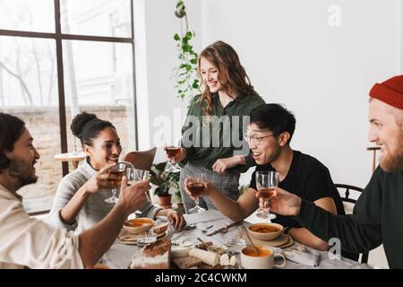 Eine Frau mit einem Lächeln und welligen Haaren hält ein Glas Wein in der Hand und steht neben dem Tisch voller Essen. Gruppe von attraktiven internationalen Freunden freudig Stockfoto