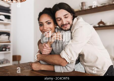 Schöner Mann, der sich traumhaft um eine schöne afroamerikanische Frau umarmt. Junge internationale Paar gerne Zeit zusammen in der gemütlichen Küche zu Hause verbringen Stockfoto