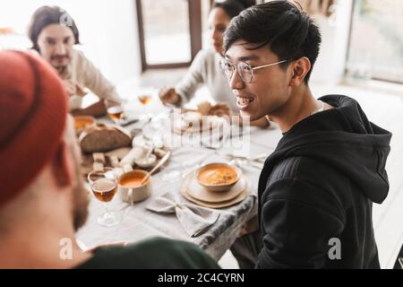 Junger lächelnder asiatischer Mann mit dunklem Haar in Brille und Kapuzenpullover, der am Tisch sitzt und glücklich zur Seite schaut. Gruppe von internationalen Freunden mit Stockfoto