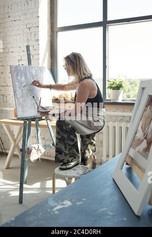 Moderne blonde Künstler in Brille mit Fuß auf Hocker und Malerei auf Leinwand mit Palette Stockfoto