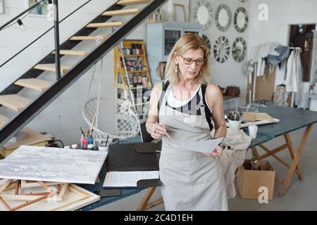 Nachdenkliche Künstlerin in Schürze und Brille beim Betrachen der Skizze während der Arbeit an einem neuen Bild im Studio Stockfoto