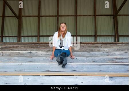 Portrait Attraktive reife Frau allein sitzen und nachdenklich auf Holztreppe oder Tribüne im Freien, in hohen Absatz Stiletto Stiefel, kopieren Raum. Stockfoto