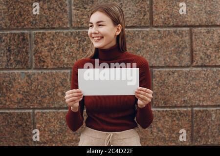 Junge Frau lächelt mit Lächeln hält weißes Papier in Hand Stein Hintergrund. Mädchen mit weißen leeren Schablone Blatt mit leerem Raum Stockfoto