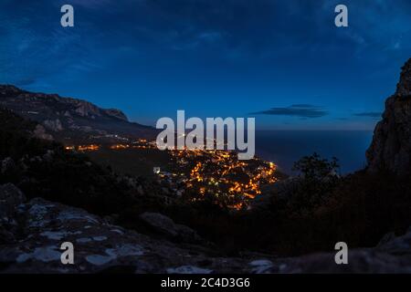 Panorama der Nachtstadt mit einer kleinen Beleuchtung des Gebäudes am Ufer des Meeres in der Nähe des Berghangs. Abendansicht der Küstenstadt Stockfoto