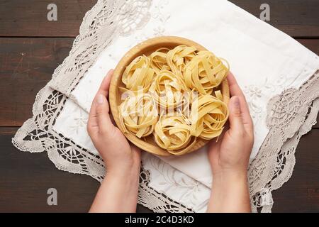 Weibliche Hände halten einen Holzteller mit roher Fettuccine Pasta über einem Holztisch, Draufsicht Stockfoto