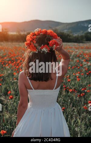 Braut in einem weißen Kleid in einem Kranz aus roten Mohnblumen, warme Sonnenuntergangszeit auf dem Hintergrund des großen roten Mohnfeldes. Speicherplatz kopieren. Das Konzept der Ruhe Stockfoto