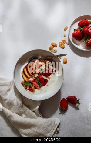 Frühstückschale mit Joghurt, Erdbeeren, Pfirsich in Scheiben geschnitten und Flocken auf einem weißen Tisch Stockfoto