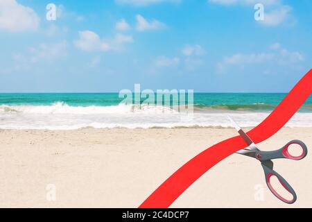 Eröffnung von Strandtourismus nach COVID-19 Quarantänekonzept. Scheren schneiden rotes Band vor dem blauen wolkigen Himmel Ozean oder Meer Sand Strand extrem nah Stockfoto