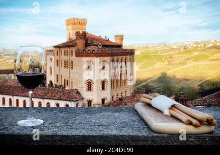 Ein Kelch des Barolo Rotweins auf einer Fensterbank mit Brotstangen und das Schloss von Barolo (Piemont, Italien) im Hintergrund Stockfoto