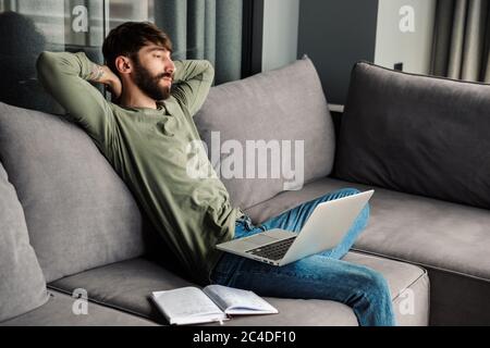 Bild von entspannten bärtigen Mann in Freizeitkleidung ruhen während der Arbeit mit Laptop auf dem Sofa im Wohnzimmer Stockfoto