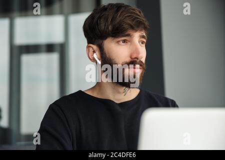 Bild eines jungen, ernsthaften Mannes, der schnurlose Ohrhörer benutzt und im Wohnzimmer mit einem Laptop arbeitet Stockfoto