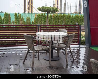 Ein Tisch und Stühle stehen auf dem nassen Holzboden eines Straßencafés vor dem Hintergrund von Bäumen und einem Gebäude im Bau Stockfoto