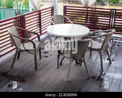 Korbsessel stehen um einen weißen runden Tisch auf einem Holzboden, der nach Regen in einem Café im Freien nass ist Stockfoto