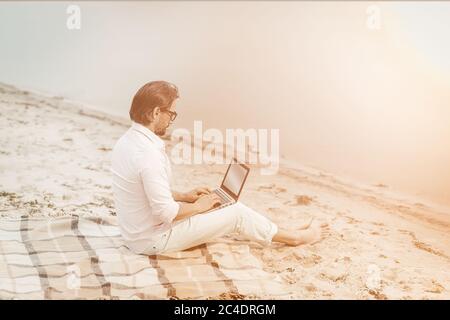Copy Writer arbeitet mit Laptop am Meer. Seitenansicht des smarten kaukasischen Geschäftsmannes, der Computer benutzt, während er auf einer Decke in der Nähe des Meeres sitzt. Getöntes Bild Stockfoto