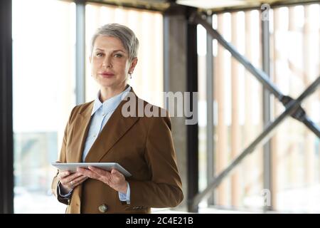 Porträt einer reifen Geschäftsfrau in formeller Kleidung, die im Büro stehend und mit einem Tablet-pc auf die Kamera schaut Stockfoto