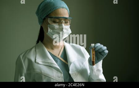 Ein medizinischer Labormitarbeiter schaut sich das Blutteströhrchen mit Covid 19-Inschrift an. Junge asiatische Frau trägt Uniform, Handschuhe und Brille auf grauem Hintergrund Stockfoto