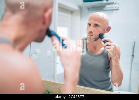 Glatze Mann Fixierung seinen Bart suchen im Badezimmerspiegel mit einem elektrischen wiederaufladbaren Bartschneider. Tägliche Körper und Hautpflege Routinebehandlungen conce Stockfoto