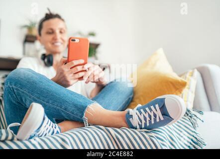 Junge Frau sitzt auf gemütlichen komfortablen Wohnzimmer Sofa, lächeln und chatten mit Freunden mit modernen Smartphone. Selektiver Fokus auf neue blaue Snea Stockfoto