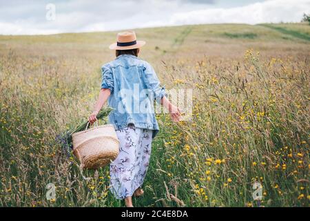 Junge Frau gekleidet Jeans Jacke und leichte Sommerkleid zu Fuß durch die hohe grüne Wiese mit Korb und Wildblumen Bouquet. Mensch und Natur c Stockfoto
