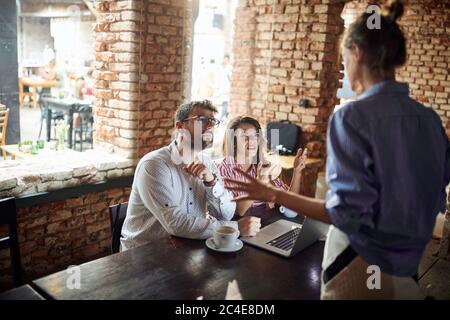 Kellnerin streitet mit einem jungen Paar im Café mit einem Laptop vor ihnen. Freiberufler Stockfoto