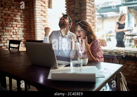 Junge kaukasische Paar Blick auf Laptop vor ihnen, Empfang gute Nachrichten, glücklich, zufrieden, Kaffee trinken im Café Stockfoto