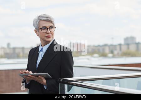 Serious reifen Geschäftsfrau in Brillen suchen weg mit nachdenklichen Blick, während mit digitalen Tablet im Freien Stockfoto