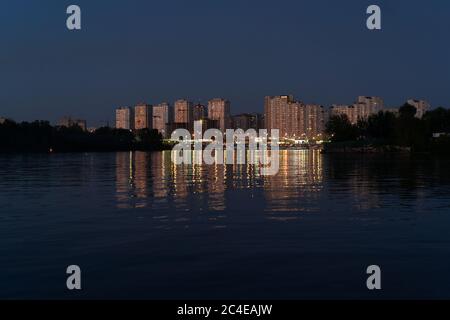 Abend am Dnipro River. Goldene Lichter moderner Gebäude spiegeln sich im Wasser. Blick auf Poznyaki Bezirk auf der linken Seite des Dnjepr. August 2019. Kiew Stockfoto