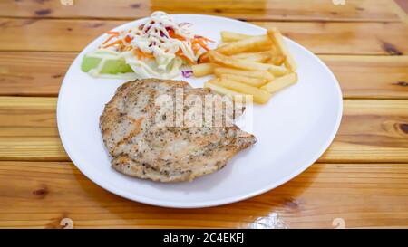 Schweinesteak mit Pommes Frites und Salat auf einem weißen Teller auf einem Holztisch. Stockfoto