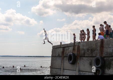 Leigh-on-Sea, Großbritannien. Juni 2020. Eine lebhafte Sommerszene am Bell Wharf Beach in Leigh-on-Sea, Essex, England. Die Menschen versammeln sich auf dem Betonpier, mit einem einzelnen Sprung ins Wasser und anderen schwimmen oder beobachten. Große Reifen säumen den Pier als Stoßfänger. Die Atmosphäre ist entspannt und verspielt und bietet spontane Küstenerholungen unter einem teilweise bewölkten Himmel. Die Leute springen von der Bell Wharf ins kühle Wasser. Leute in Old Leigh, Essex, genießen das warme Wetter und weniger überfüllte Strände. Penelope Barritt/Alamy Live News Stockfoto