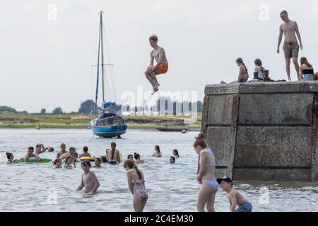 Leigh-on-Sea, Großbritannien. Juni 2020. Eine lebhafte Sommerszene am Bell Wharf Beach in Leigh-on-Sea, Essex, wo Menschen schwimmen, vom Pier springen und das Wasser genießen. Ein Segelboot liegt vor der Küste vor Anker, und die Küste ist mit grasbewachsenen Flächen und Bäumen versehen, was eine entspannte Küstenatmosphäre hervorruft. Die Leute springen von der Bell Wharf ins kühle Wasser. Leute in Old Leigh, Essex, genießen das warme Wetter und weniger überfüllte Strände. Penelope Barritt/Alamy Live News Stockfoto
