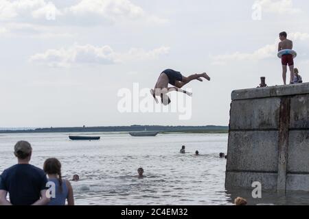 Leigh-on-Sea, Großbritannien. Juni 2020. Eine Gruppe von Menschen genießt einen lebhaften Sommernachmittag am Bell Wharf Beach in Leigh-on-Sea, Essex. Eine Person führt einen Backflip von einer Betonplattform ins Wasser aus, während andere schwimmen, beobachten und sich entlang der Küste entspannen. Boote und eine ferne Küste runden die lebendige Küstenszene ab. Die Leute springen von der Bell Wharf ins kühle Wasser. Leute in Old Leigh, Essex, genießen das warme Wetter und weniger überfüllte Strände. Penelope Barritt/Alamy Live News Stockfoto