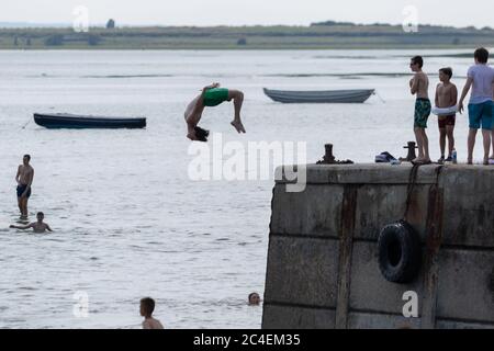 Leigh-on-Sea, Großbritannien. Juni 2020. Ein junger Mensch führt einen Backflip vom Betonpier am Bell Wharf Beach, Leigh-on-Sea, durch, während andere beobachten oder sich darauf vorbereiten zu springen. Die Schwimmer genießen das Wasser unter sich, mit kleinen Booten und der Mündungslandschaft im Hintergrund. Eine lebhafte Sommerszene, in der spontane Küstenerholungen aufgenommen werden. Die Leute springen von der Bell Wharf ins kühle Wasser. Leute in Old Leigh, Essex, genießen das warme Wetter und weniger überfüllte Strände. Penelope Barritt/Alamy Live News Stockfoto