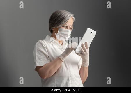 Seitenansicht des Arztes mit elektronischer Tablette. Kaukasische gealterte Frau in Schutzmaske arbeitet mit digitalen Gadget Blick auf Bildschirm auf graue Wand Stockfoto